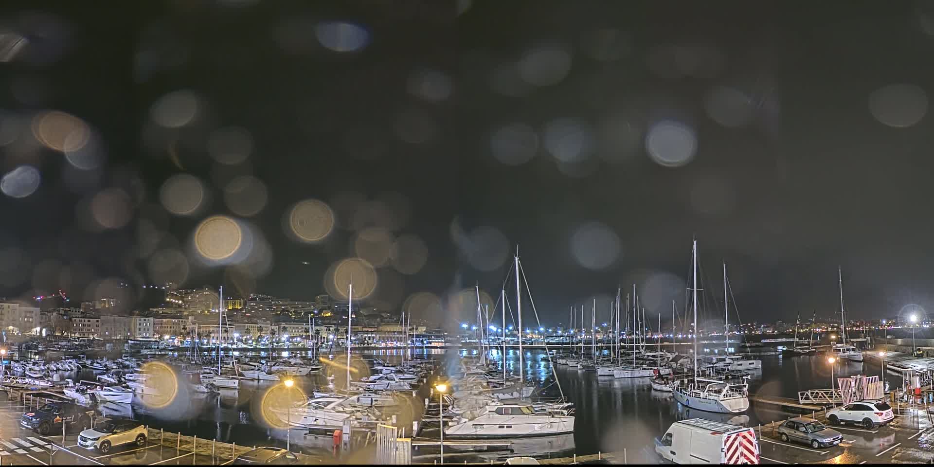 A nighttime view of a marina filled with boats, seen through rain-covered glass, with a city skyline in the background.