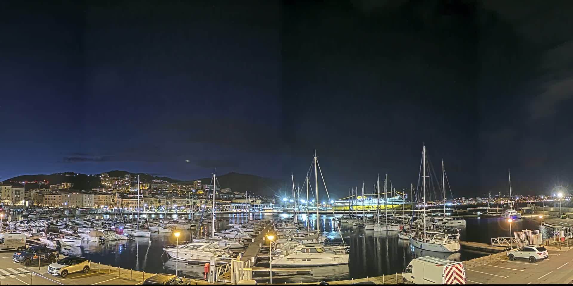 A nighttime view of a marina filled with sailboats and motorboats, with a city skyline and hills in the background under a clear, dark sky.