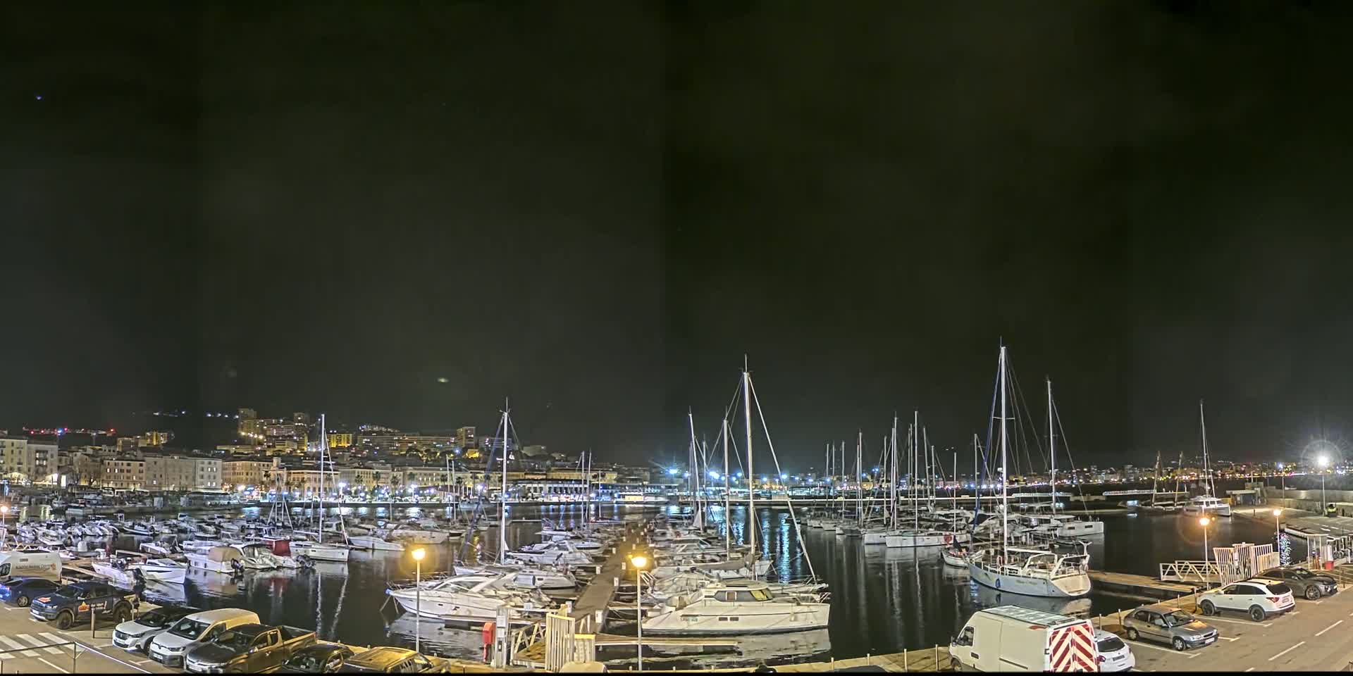 A nighttime view of a marina filled with numerous boats, docked alongside a city illuminated with lights under a dark, clear sky.