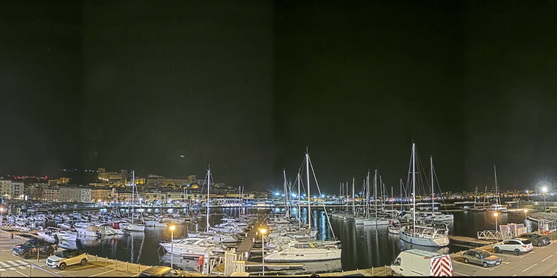 A nighttime view of a marina filled with sailboats and motorboats, under a clear, dark sky, with a city skyline visible in the distance.