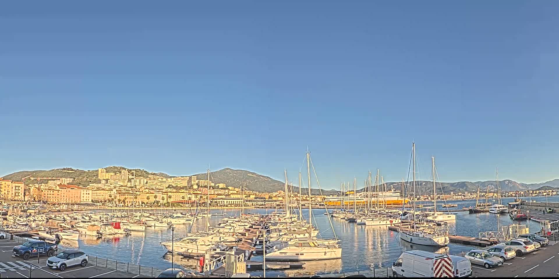 A harbor filled with numerous sailboats and motorboats is situated in front of a city nestled against mountains under a clear blue sky.