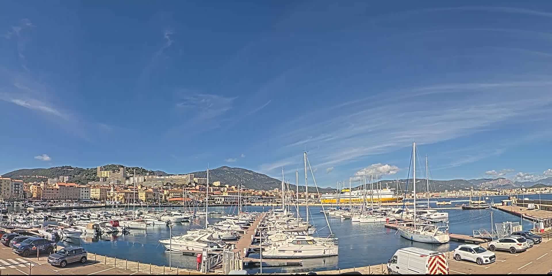 A marina filled with sailboats and motorboats is situated in front of a city nestled against mountains under a mostly sunny sky with a few scattered clouds.