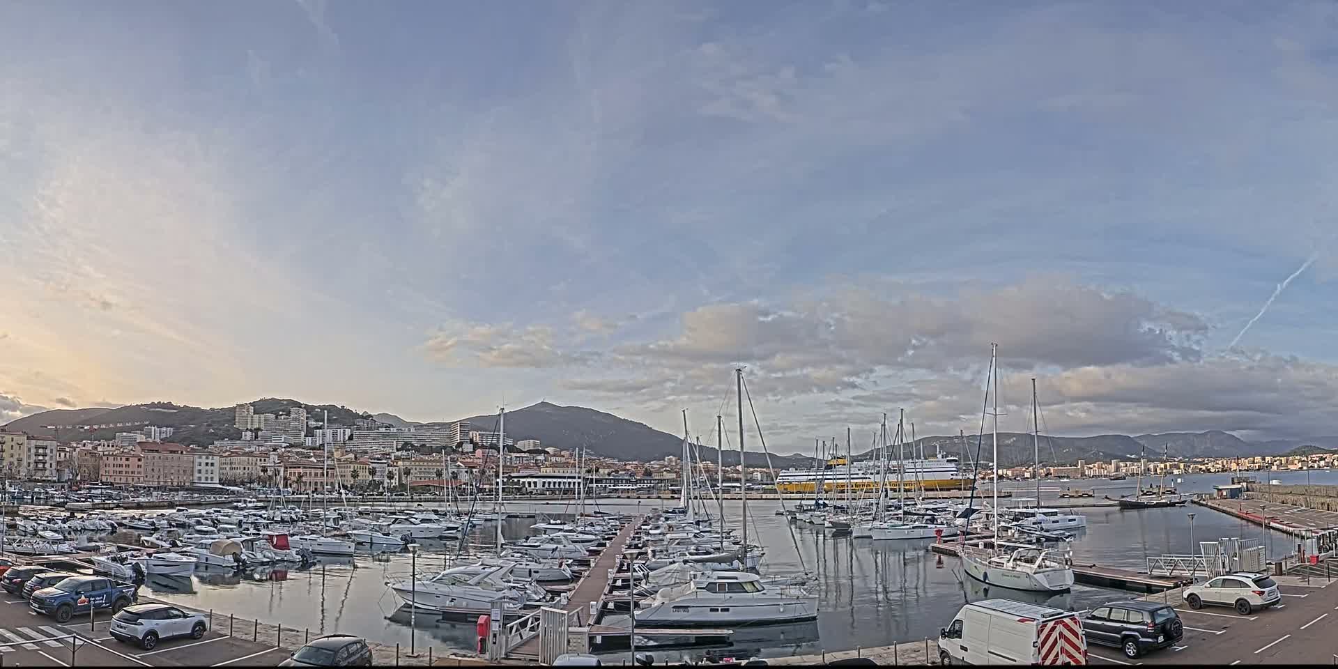 A marina filled with sailboats and motorboats is situated in front of a city nestled against a mountain range under a mostly cloudy sky.