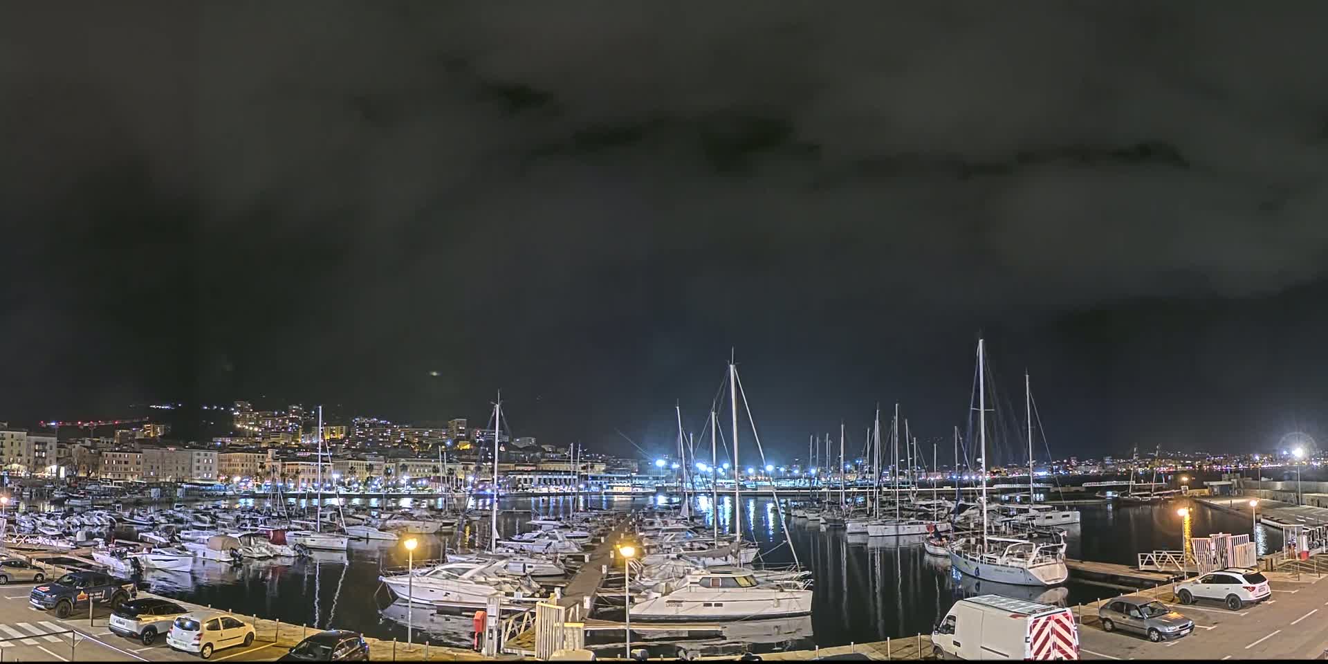 A marina filled with numerous boats is illuminated at night under a dark, cloudy sky.