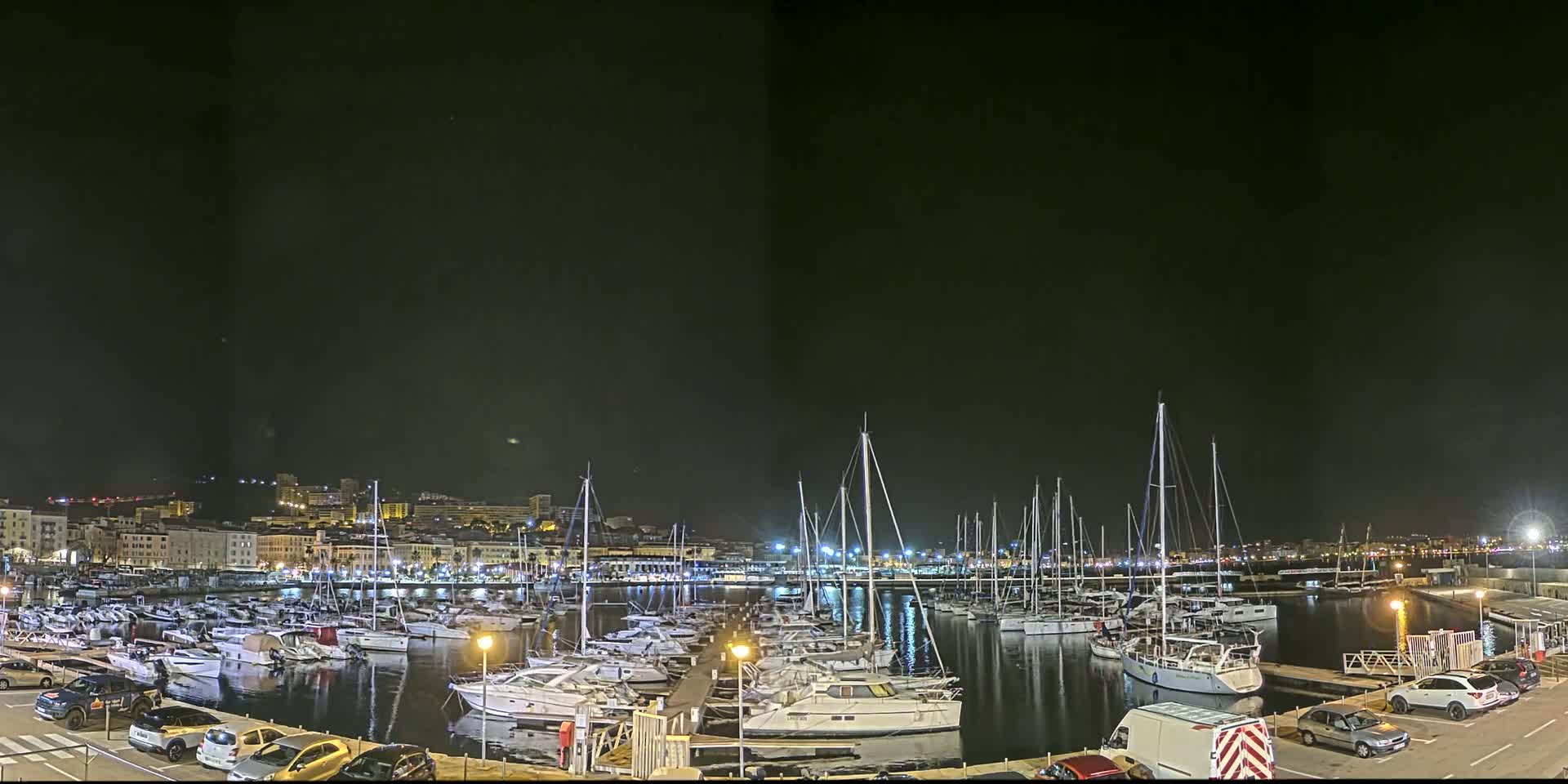 A marina filled with sailboats and motorboats is illuminated at night, with a city skyline in the background under a clear, dark sky.