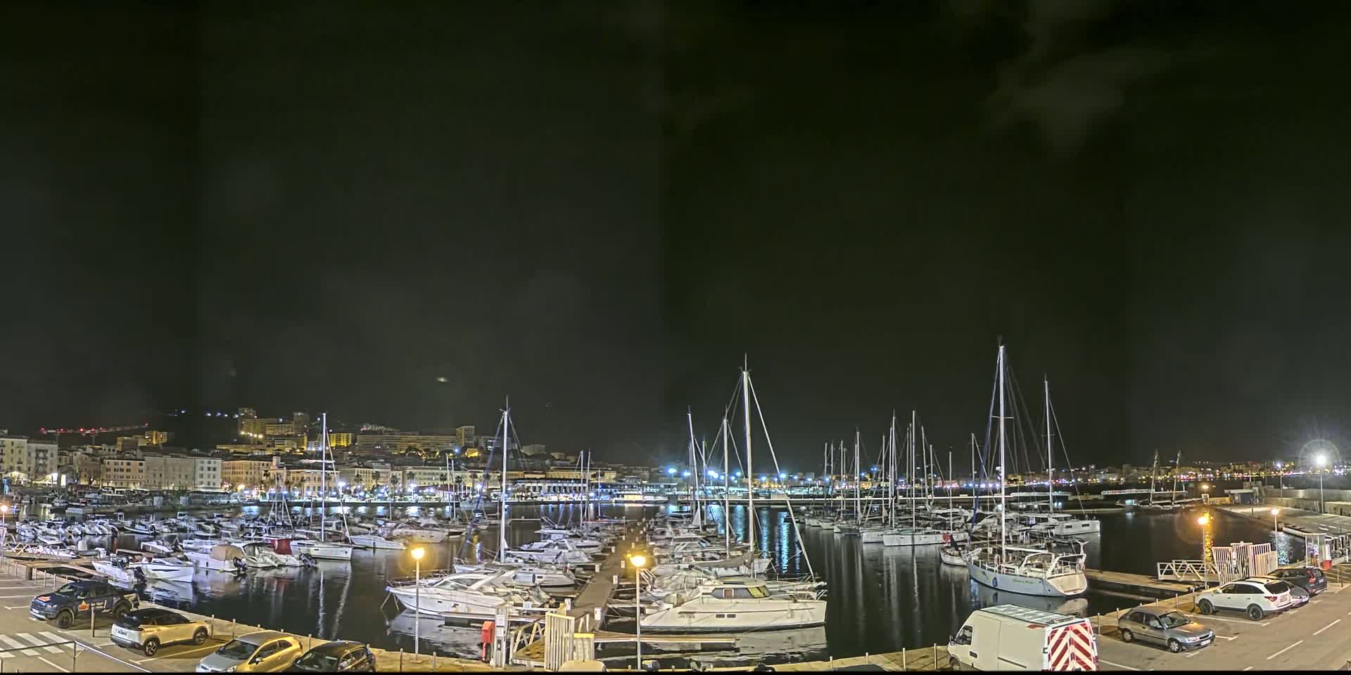 A nighttime view of a marina filled with numerous boats and yachts, with a city skyline illuminated in the background under a dark, cloudy sky.