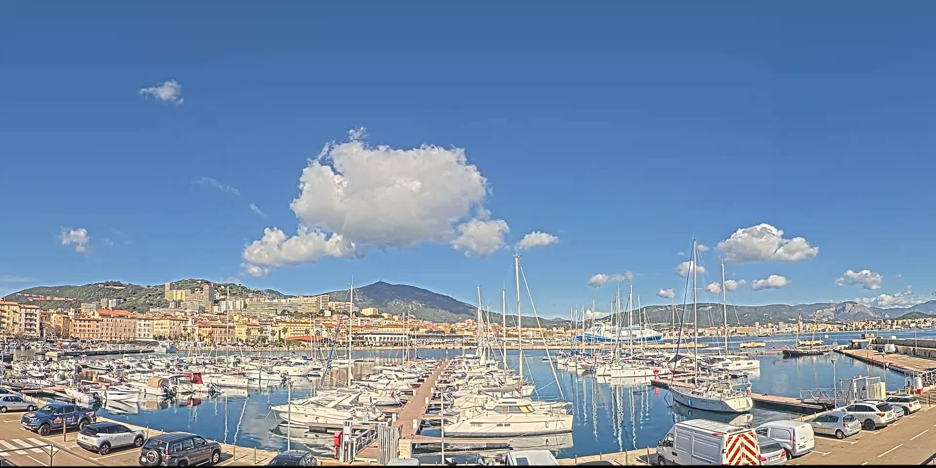 A marina filled with sailboats and motorboats is situated in front of a city, all under a mostly sunny sky with a few scattered clouds.