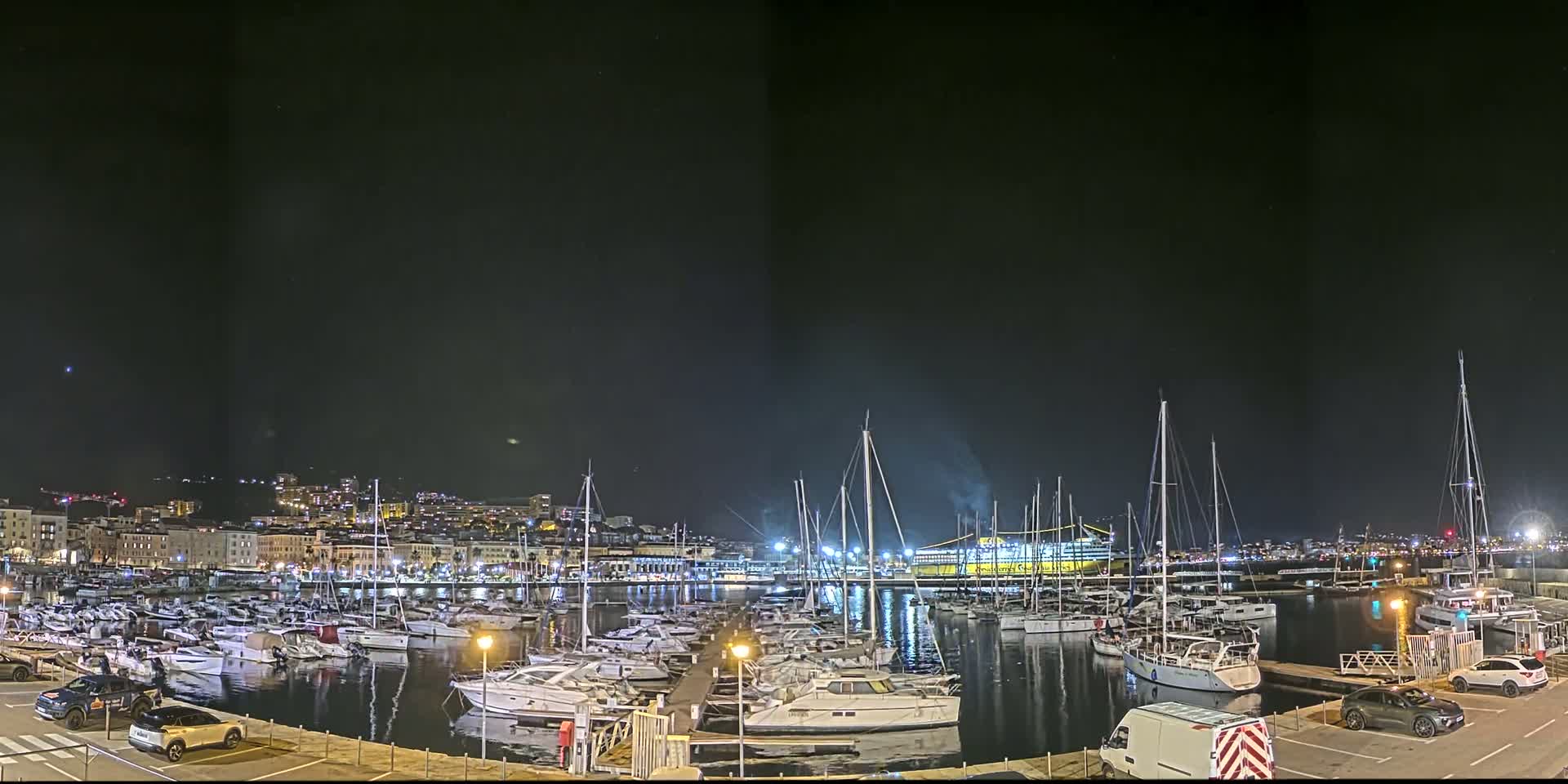 A nighttime view of a harbor filled with numerous boats and yachts, with a city skyline visible in the background under a clear, dark sky.