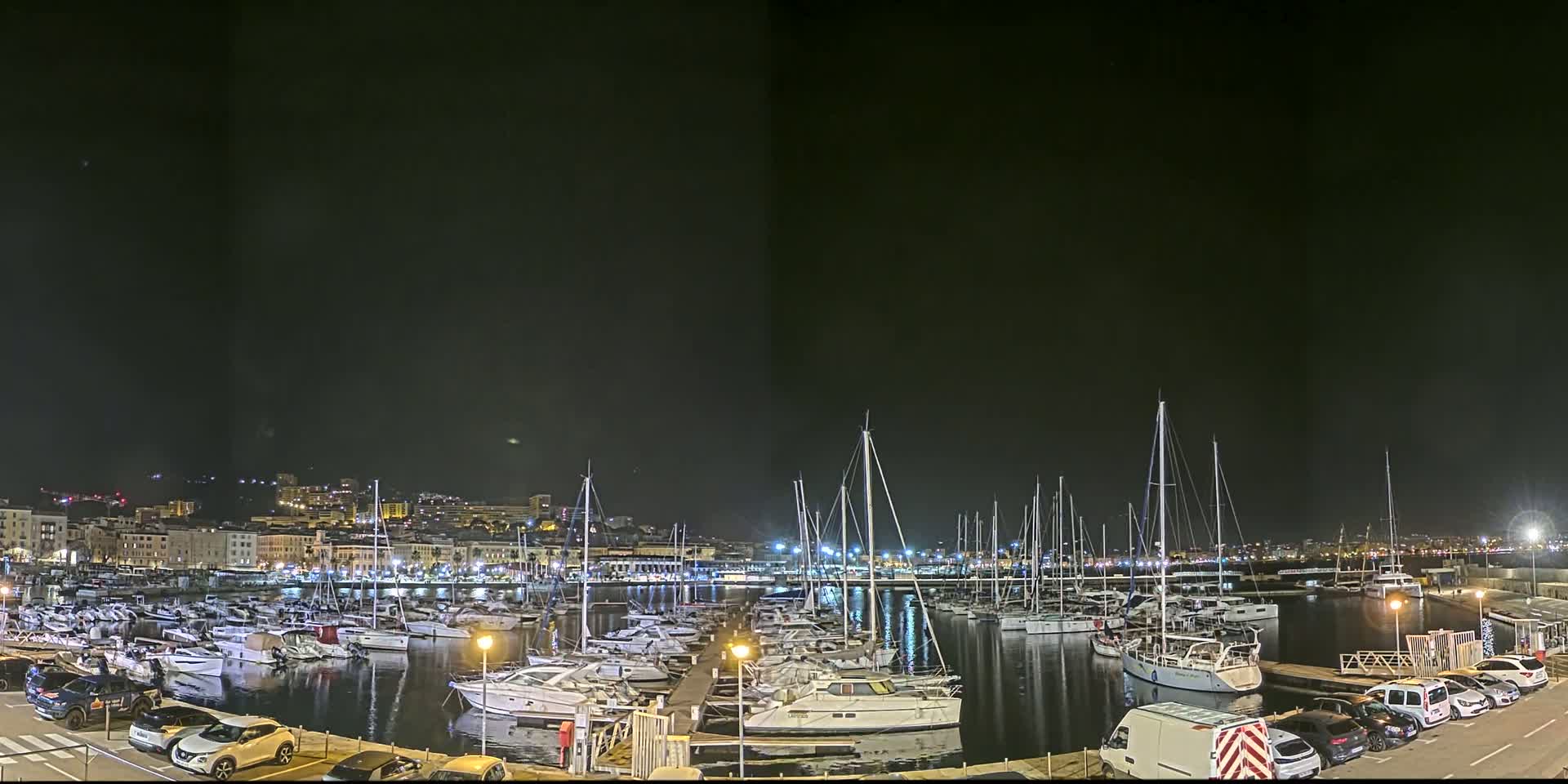 A nighttime view of a marina filled with boats, with a city skyline illuminated in the background under a clear, dark sky.