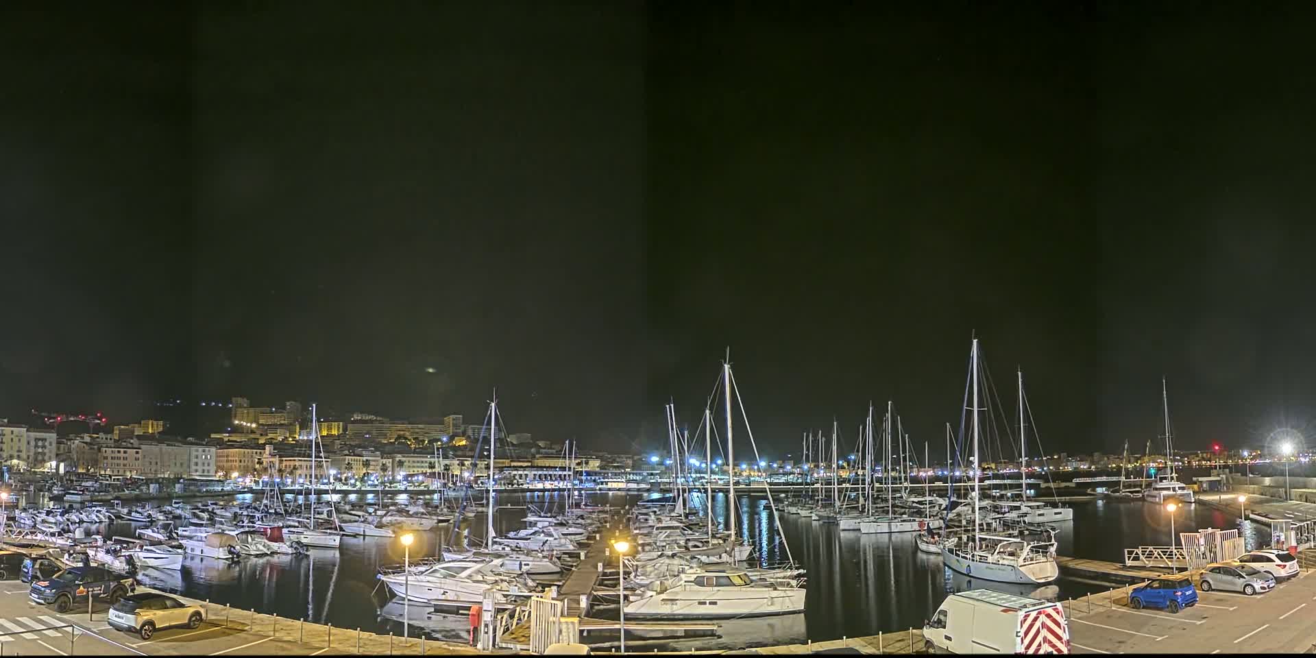 A nighttime view of a marina filled with numerous sailboats and motorboats, docked along a waterfront with a city skyline in the background.