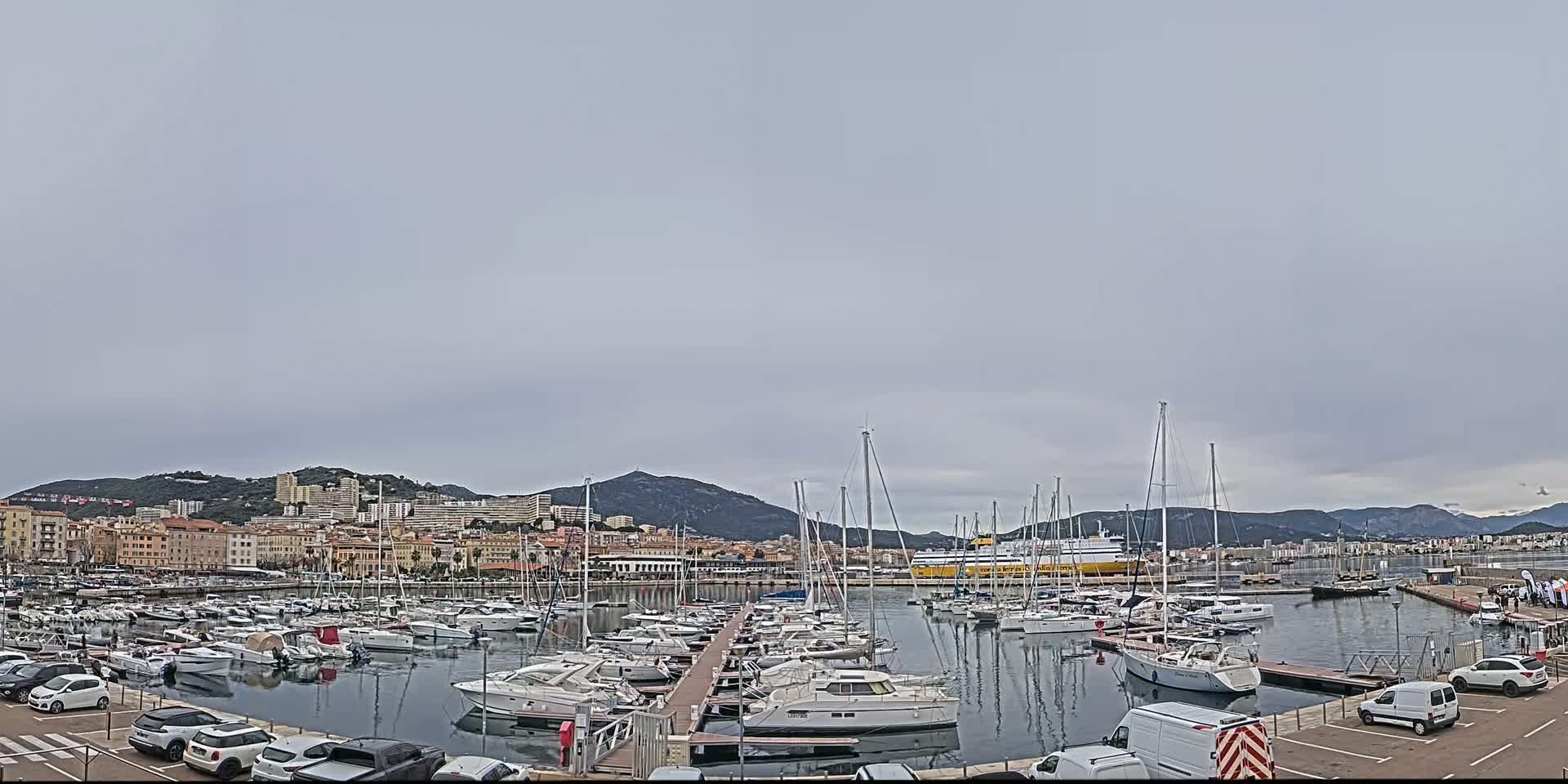 A harbor filled with numerous sailboats and motorboats is situated in front of a city nestled against a mountain range under an overcast sky.