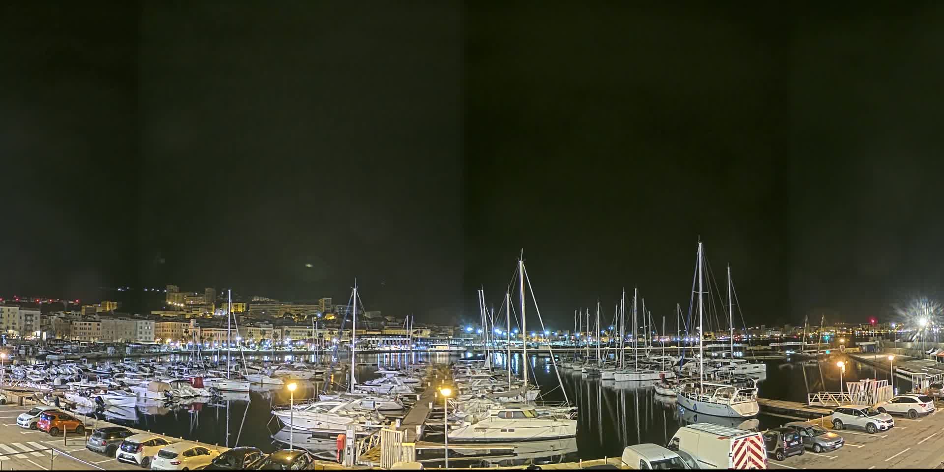 A marina filled with numerous boats is illuminated at night, with a city skyline visible in the background under a clear, dark sky.