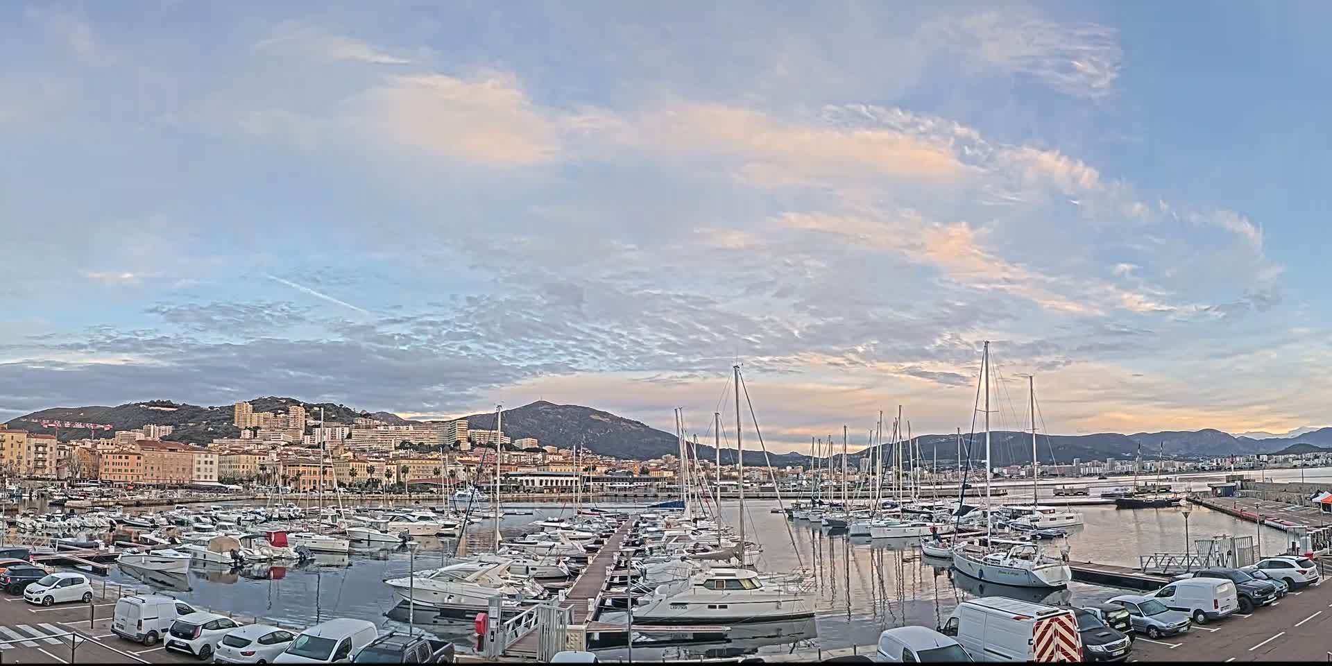 A marina filled with numerous sailboats and motorboats is situated in front of a city nestled against a mountain range under a partly cloudy sky.