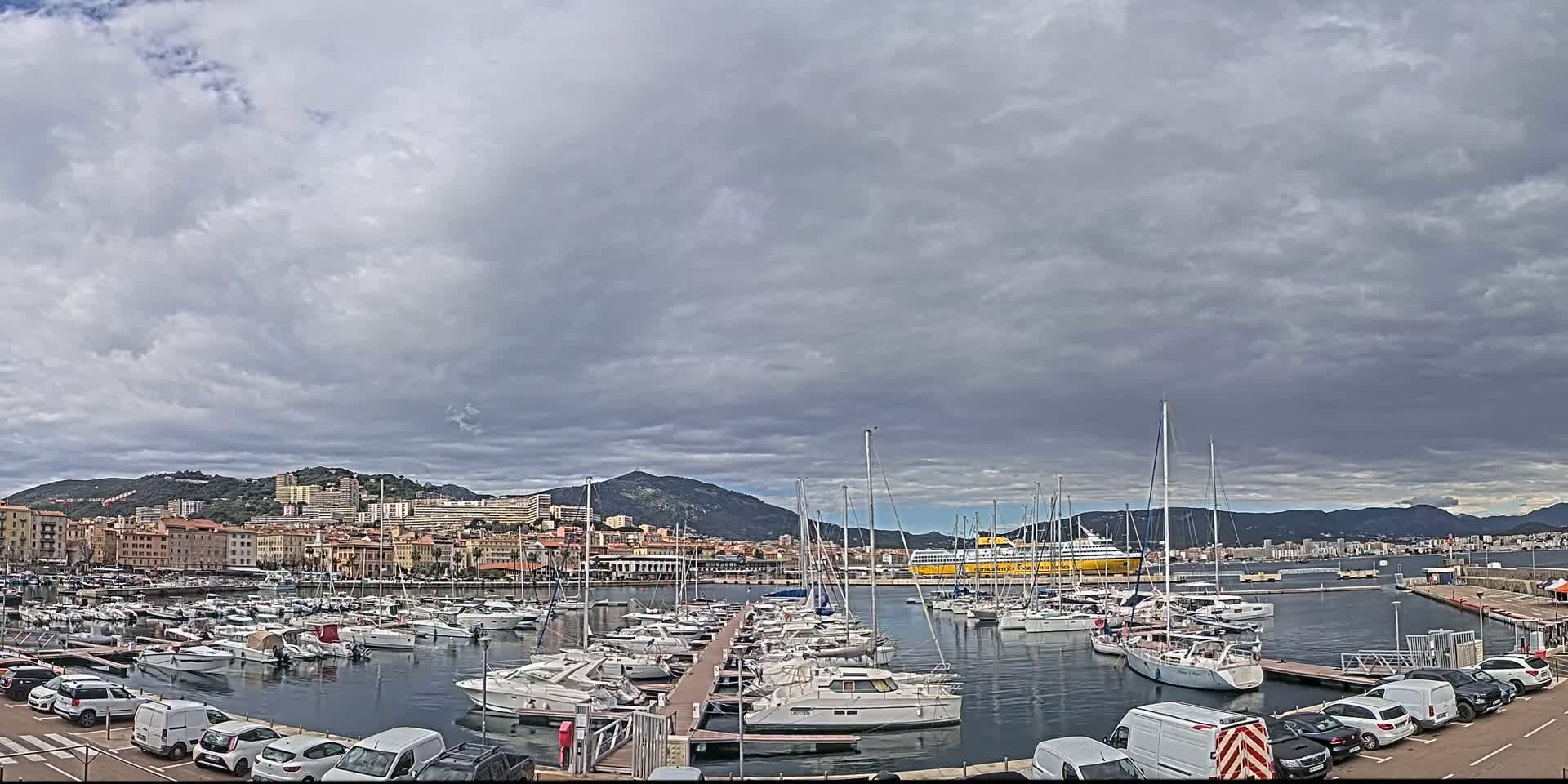 A harbor filled with sailboats and yachts is situated in front of a city under a mostly cloudy sky.