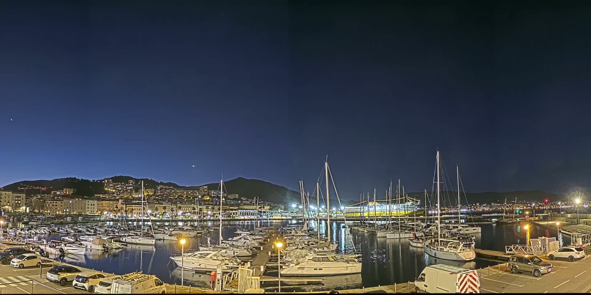 A nighttime view of a marina filled with boats, docked along a waterfront city under a clear, dark blue sky.