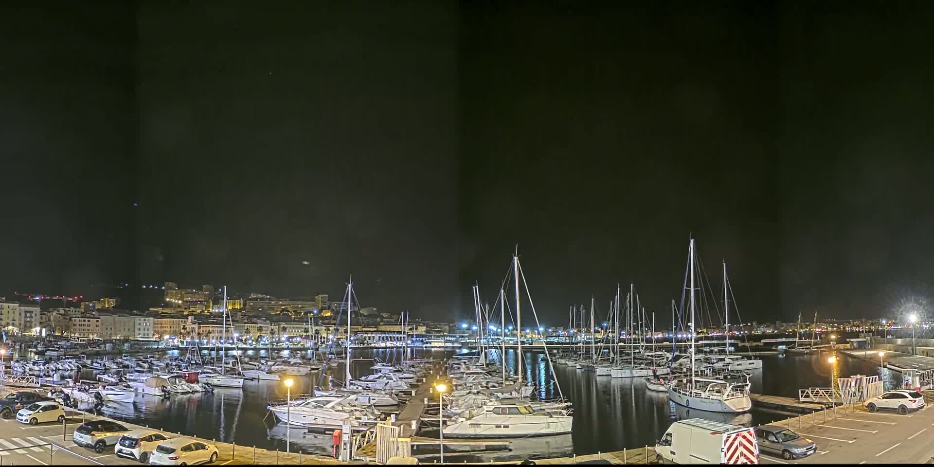 A marina filled with boats is illuminated at night under a clear, dark sky, with a city skyline visible in the background.