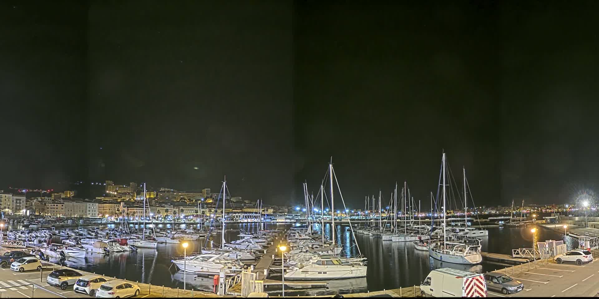 A marina filled with sailboats and motorboats is illuminated at night under a clear, dark sky, with a city skyline visible in the background.