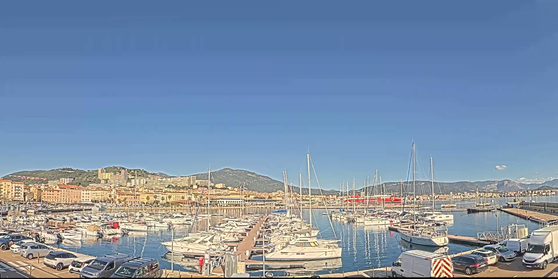 A harbor filled with numerous sailboats and motorboats is nestled beside a city against a backdrop of mountains under a clear blue sky.