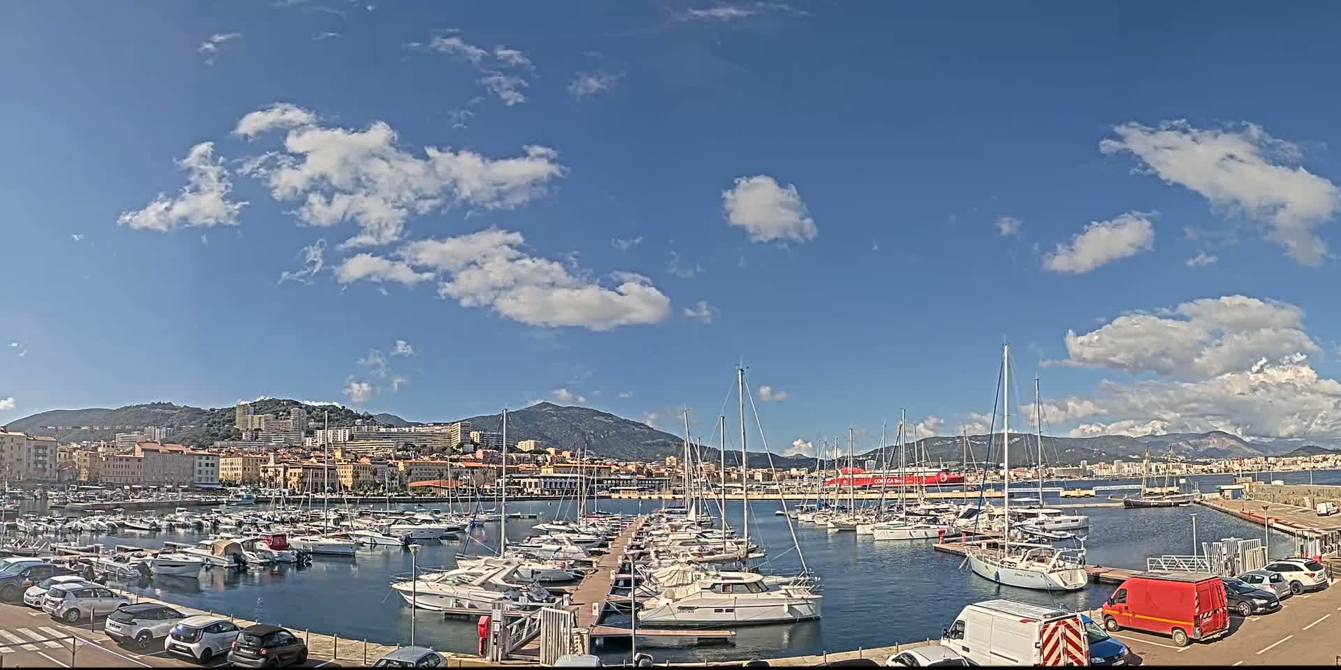 A harbor filled with sailboats and motorboats is situated in front of a city nestled against a mountain range under a mostly sunny sky with scattered clouds.