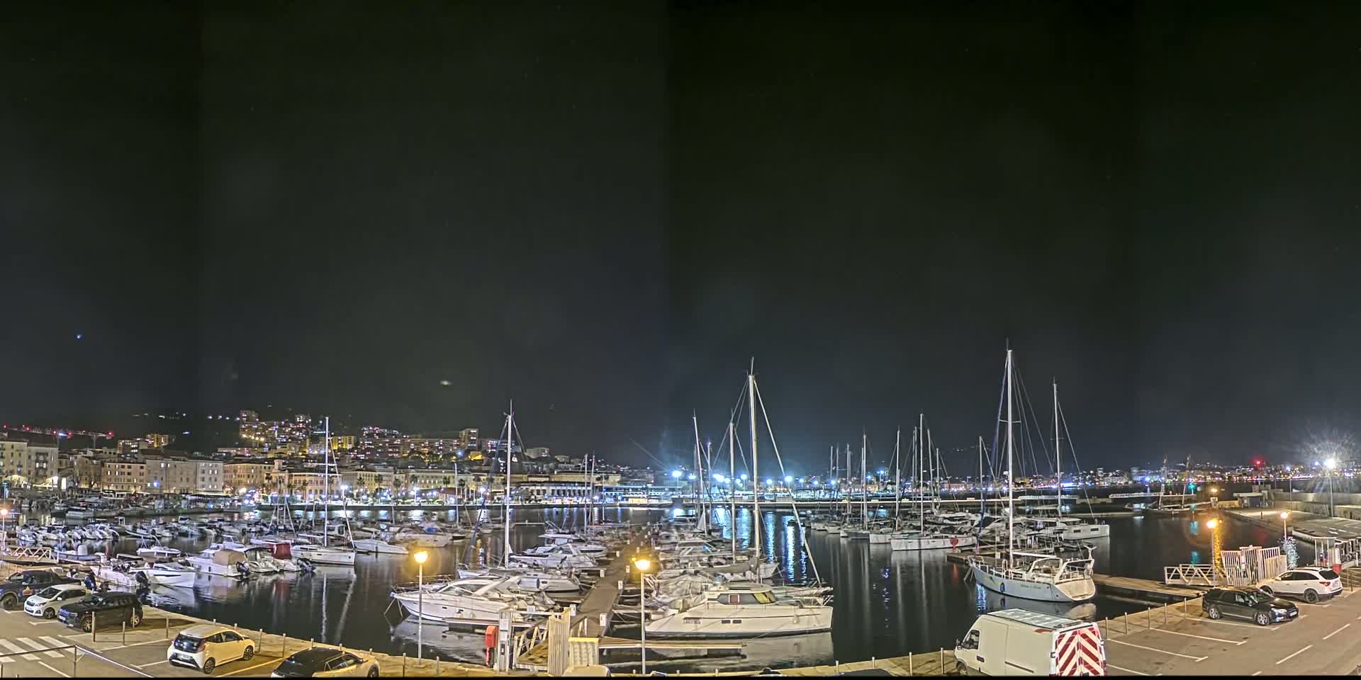 A marina filled with numerous boats is illuminated at night under a clear, dark sky, with a city skyline visible in the background.