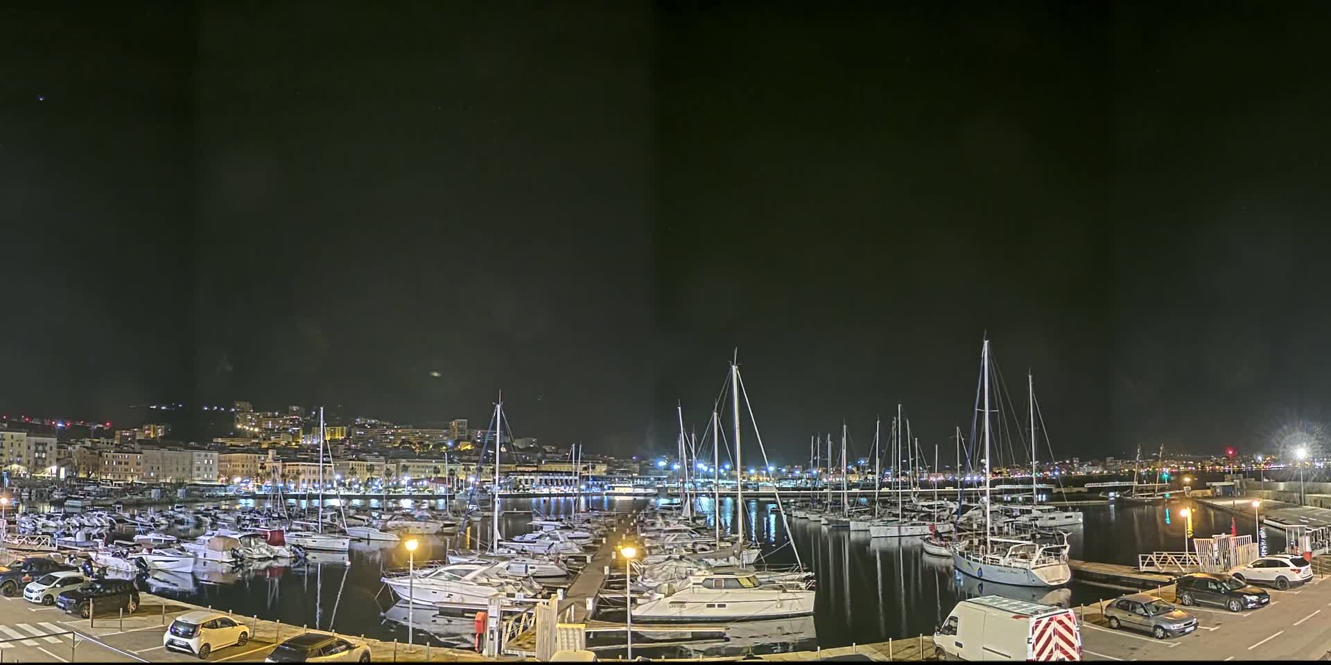 A nighttime view of a marina filled with numerous boats, docked alongside a city illuminated by lights under a clear, dark sky.
