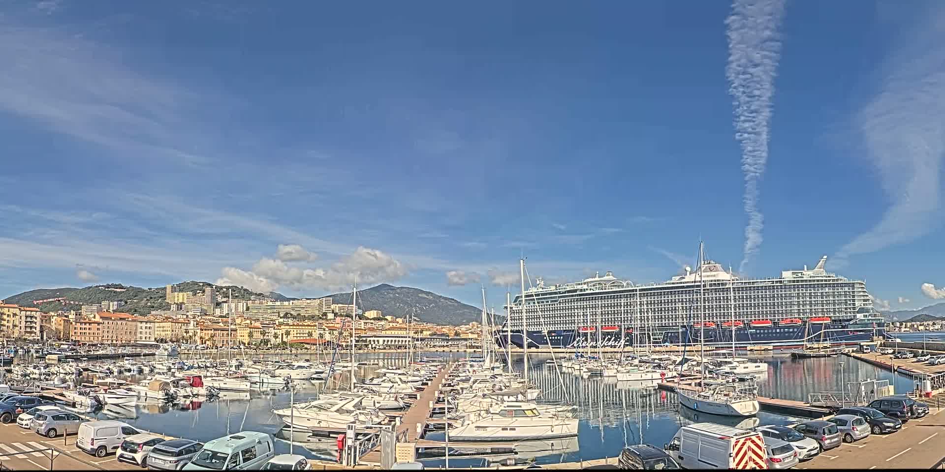 A large cruise ship is docked in a harbor filled with numerous smaller boats, under a mostly sunny sky with some thin clouds and a contrail.