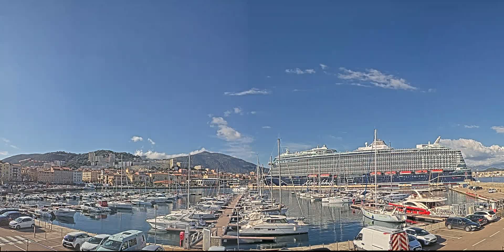 A large cruise ship is docked in a harbor filled with smaller boats, under a mostly clear blue sky with a few scattered clouds, and backed by a cityscape nestled against hills.