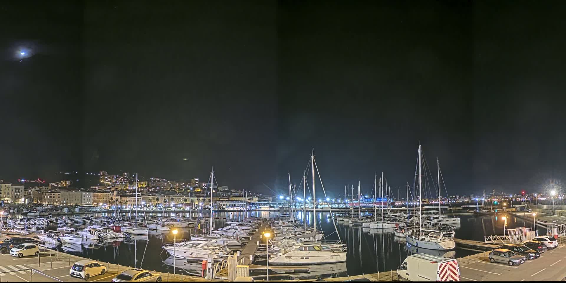 A nighttime view of a marina filled with sailboats and motorboats, illuminated by city lights under a dark sky.