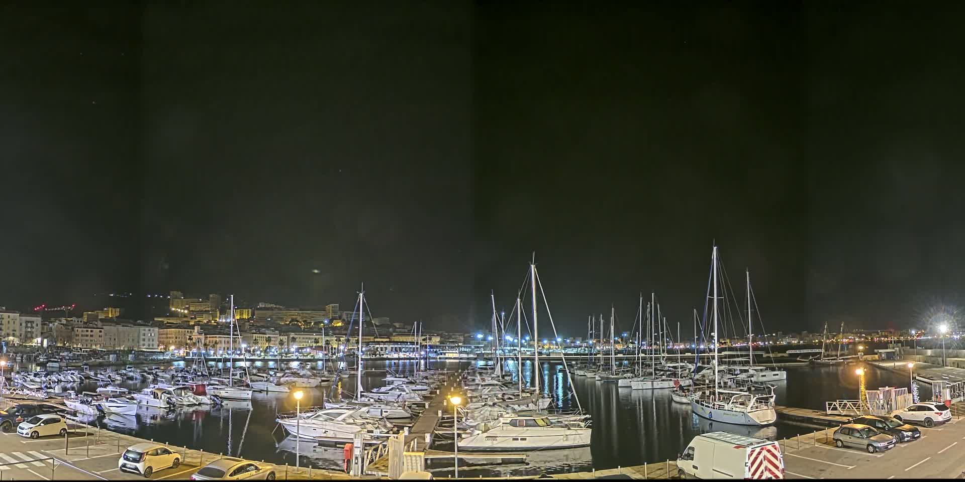 A nighttime view of a harbor filled with numerous boats, docked alongside a city illuminated by lights under a clear, dark sky.
