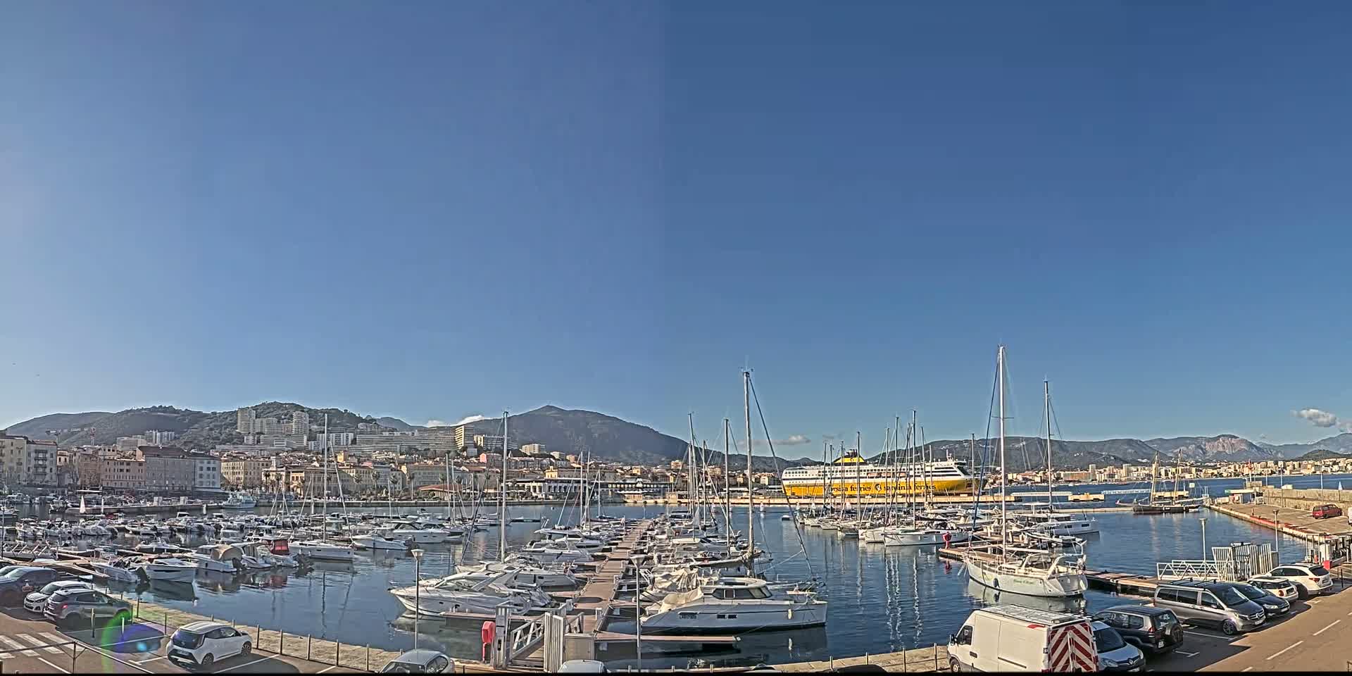 A marina filled with numerous sailboats and motorboats is situated in front of a city skyline and mountains under a clear, bright blue sky.