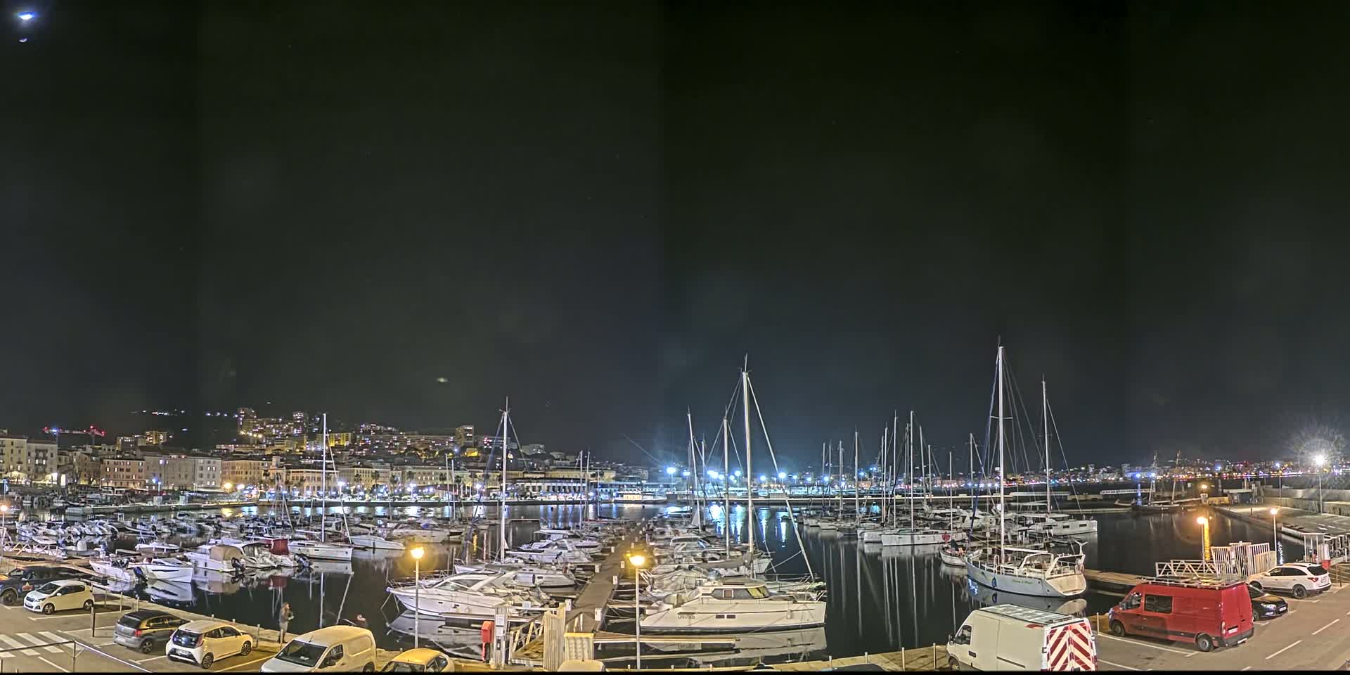 A marina filled with docked sailboats and motorboats is illuminated at night under a clear, moonlit sky.