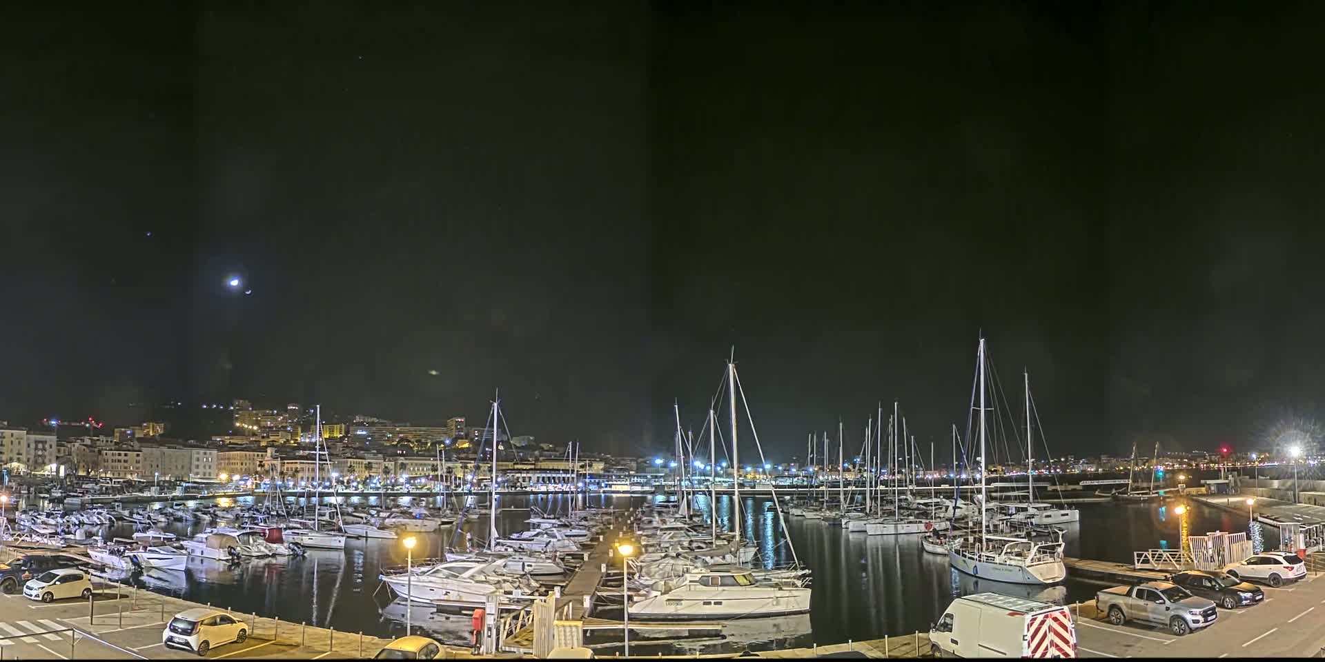 A nighttime harbor filled with many docked sailboats and motorboats under a clear, dark sky with a visible moon.