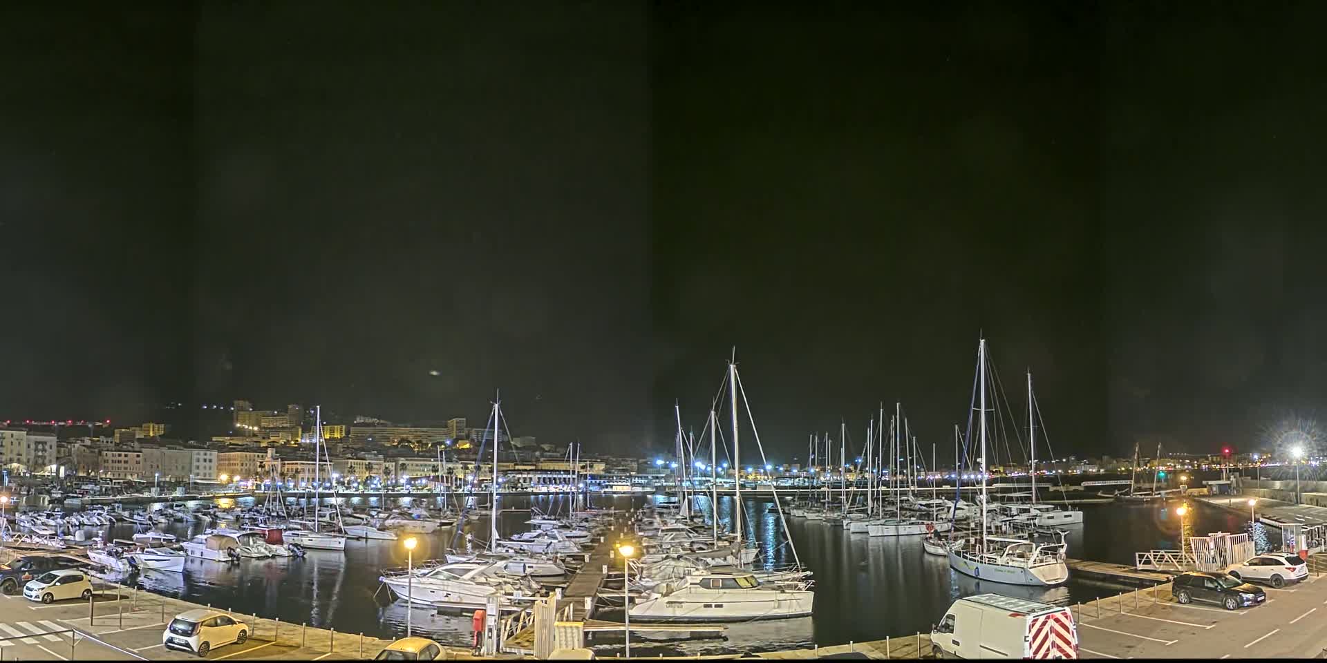 A nighttime view of a marina filled with numerous boats docked alongside a city's illuminated waterfront under a dark, clear sky.