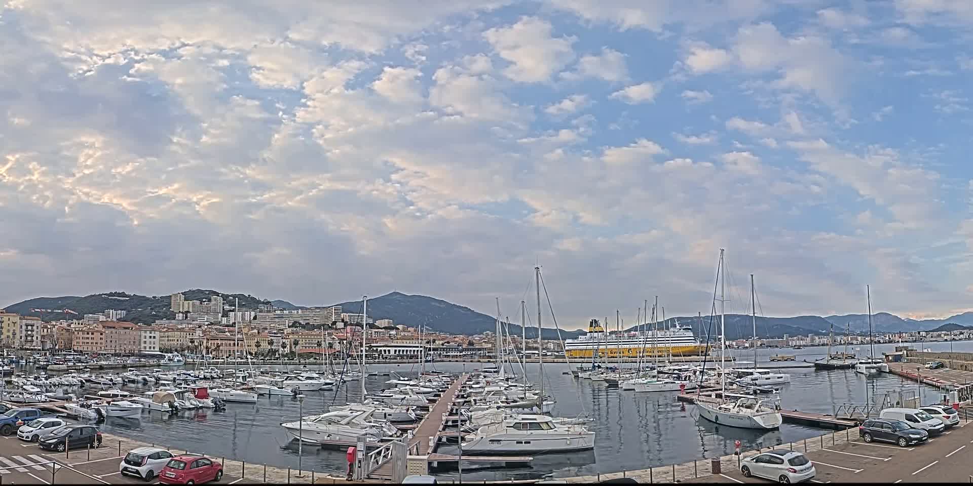 A partly cloudy day overlooks a marina filled with numerous boats and yachts, a large ferry in the distance, and a city in the background with mountains beyond.