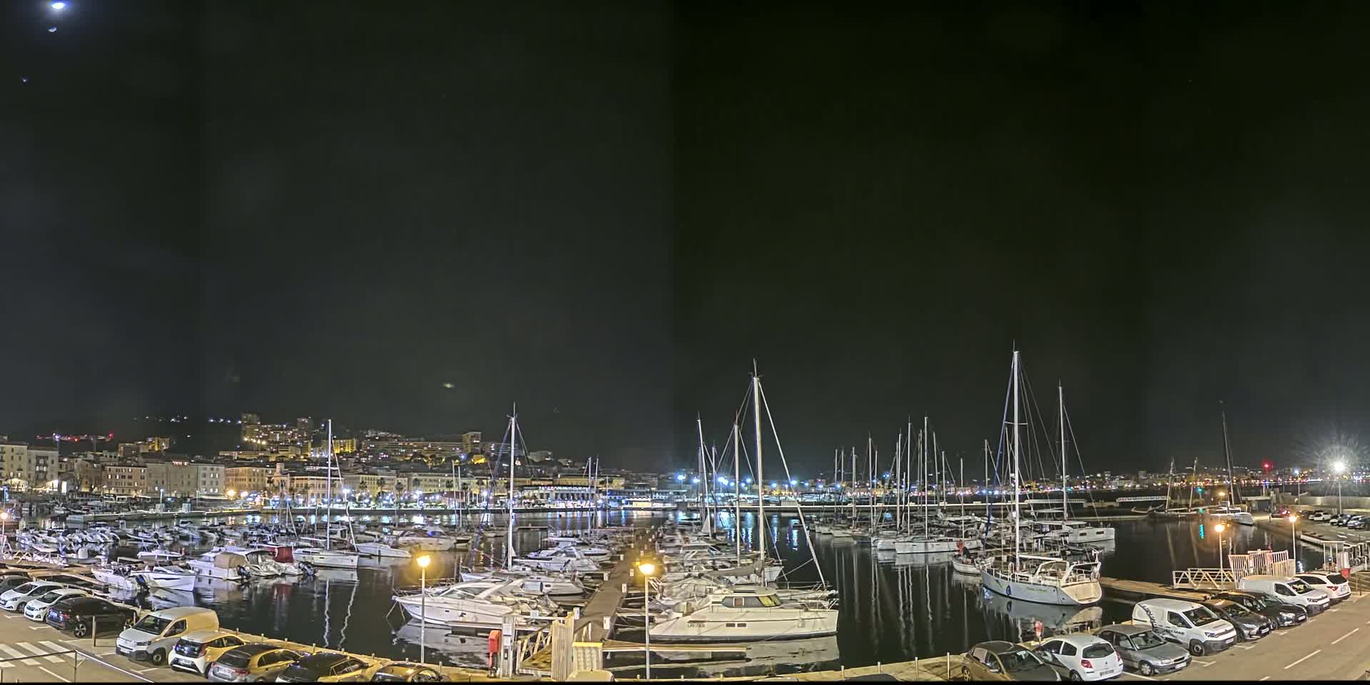 A nighttime harbor filled with numerous boats and yachts is illuminated under a mostly clear sky with a visible moon.