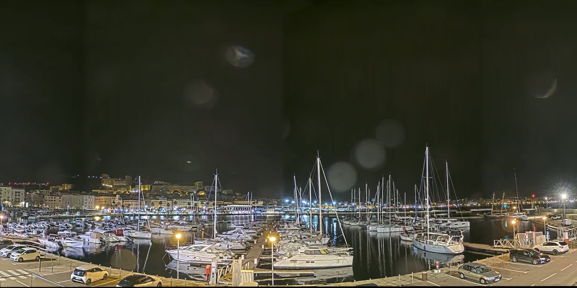 A nighttime view of a marina filled with numerous boats docked along a waterfront, under a dark, slightly rainy sky.