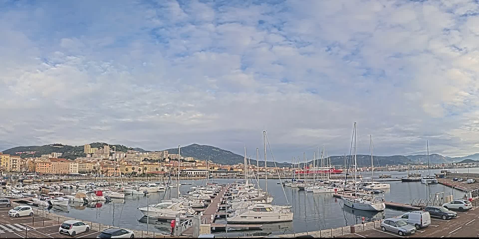 A harbor filled with many sailboats and motorboats is situated in front of a city nestled against mountains under a partly cloudy sky.