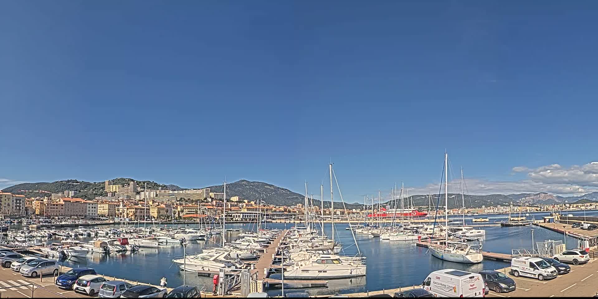 A marina filled with numerous sailboats and motorboats is situated in front of a city nestled against a mountain range under a clear blue sky.