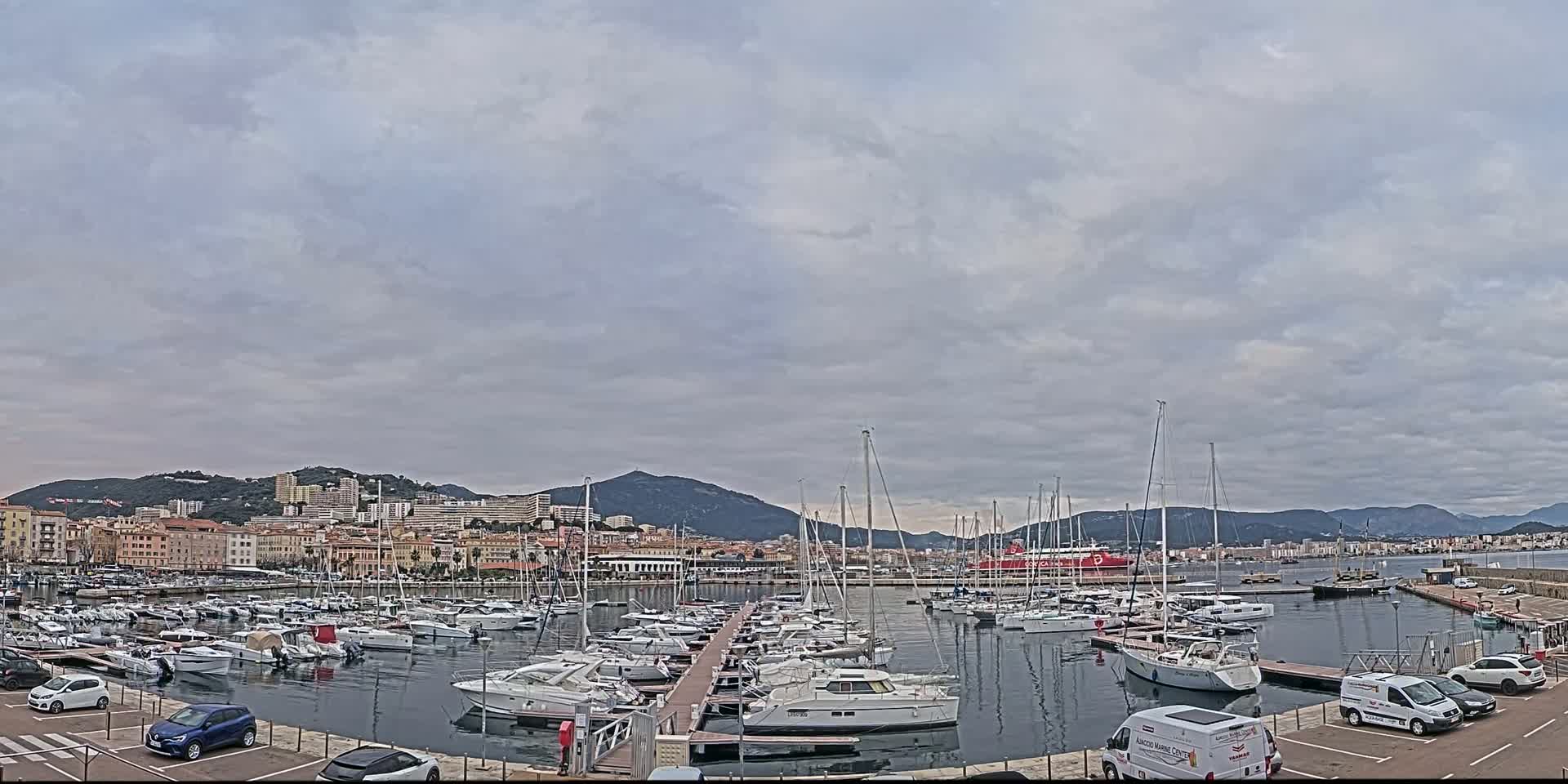 A marina filled with sailboats and yachts is situated in front of a city nestled against mountains under a cloudy sky.