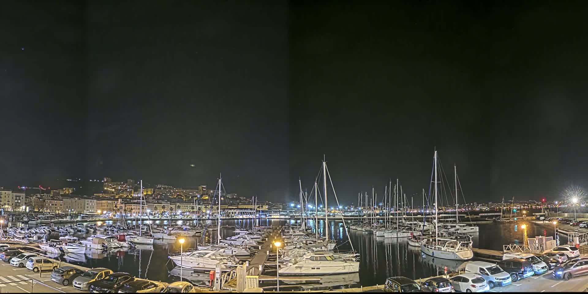 A nighttime view of a harbor filled with numerous boats and yachts, with a city skyline visible in the background under a clear, dark sky.
