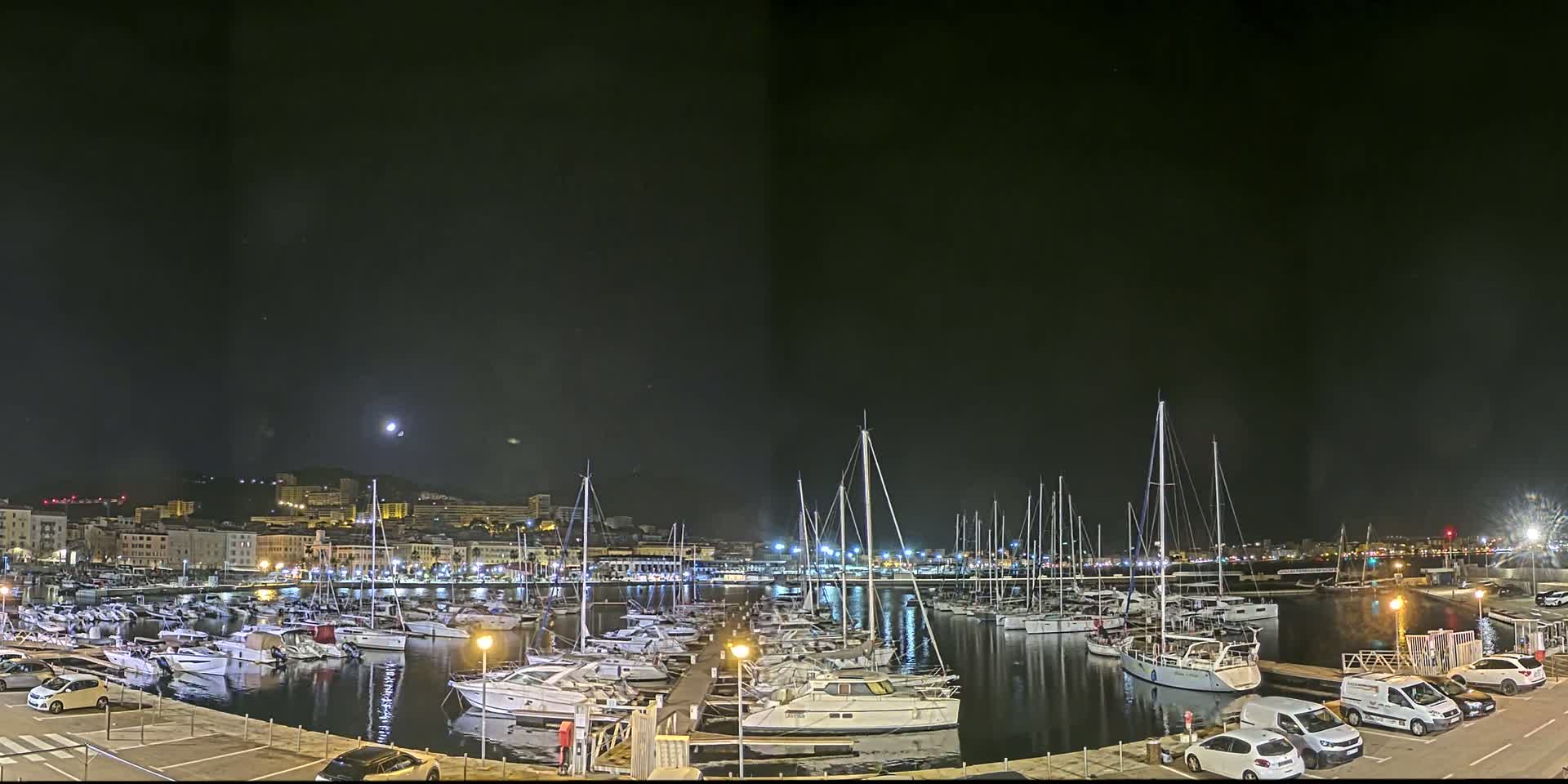 A nighttime view of a marina filled with docked sailboats and motorboats, under a mostly clear sky with a visible moon, and a city skyline in the background.