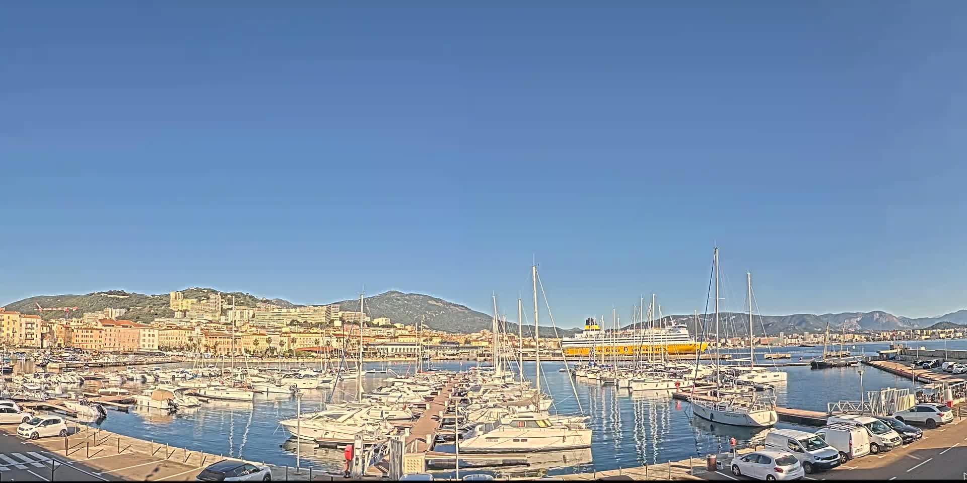 A harbor filled with sailboats and yachts is situated in front of a city and mountains under a clear blue sky.