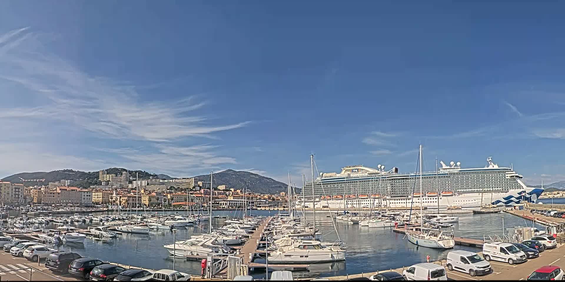 A large cruise ship is docked in a harbor filled with many smaller boats, under a mostly clear blue sky with wispy clouds.