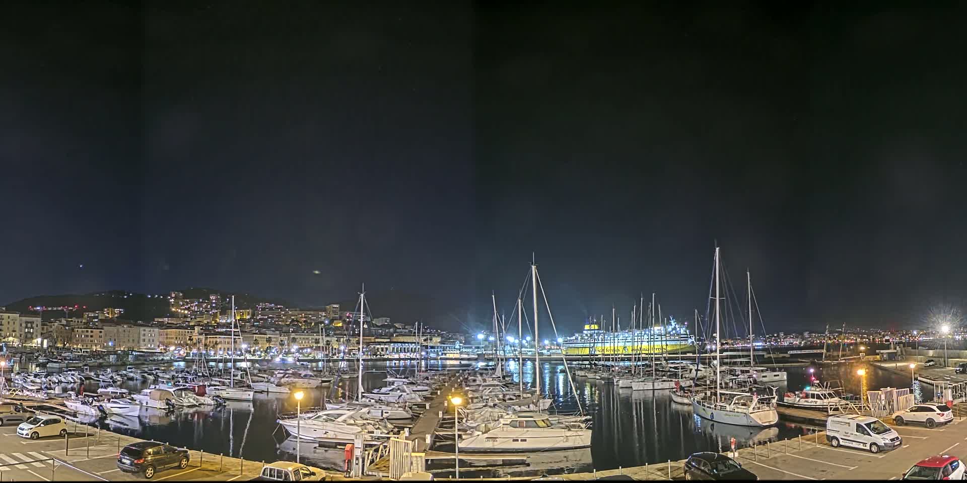 A marina filled with numerous boats is illuminated at night under a clear, dark sky, with a city skyline and hills visible in the background.