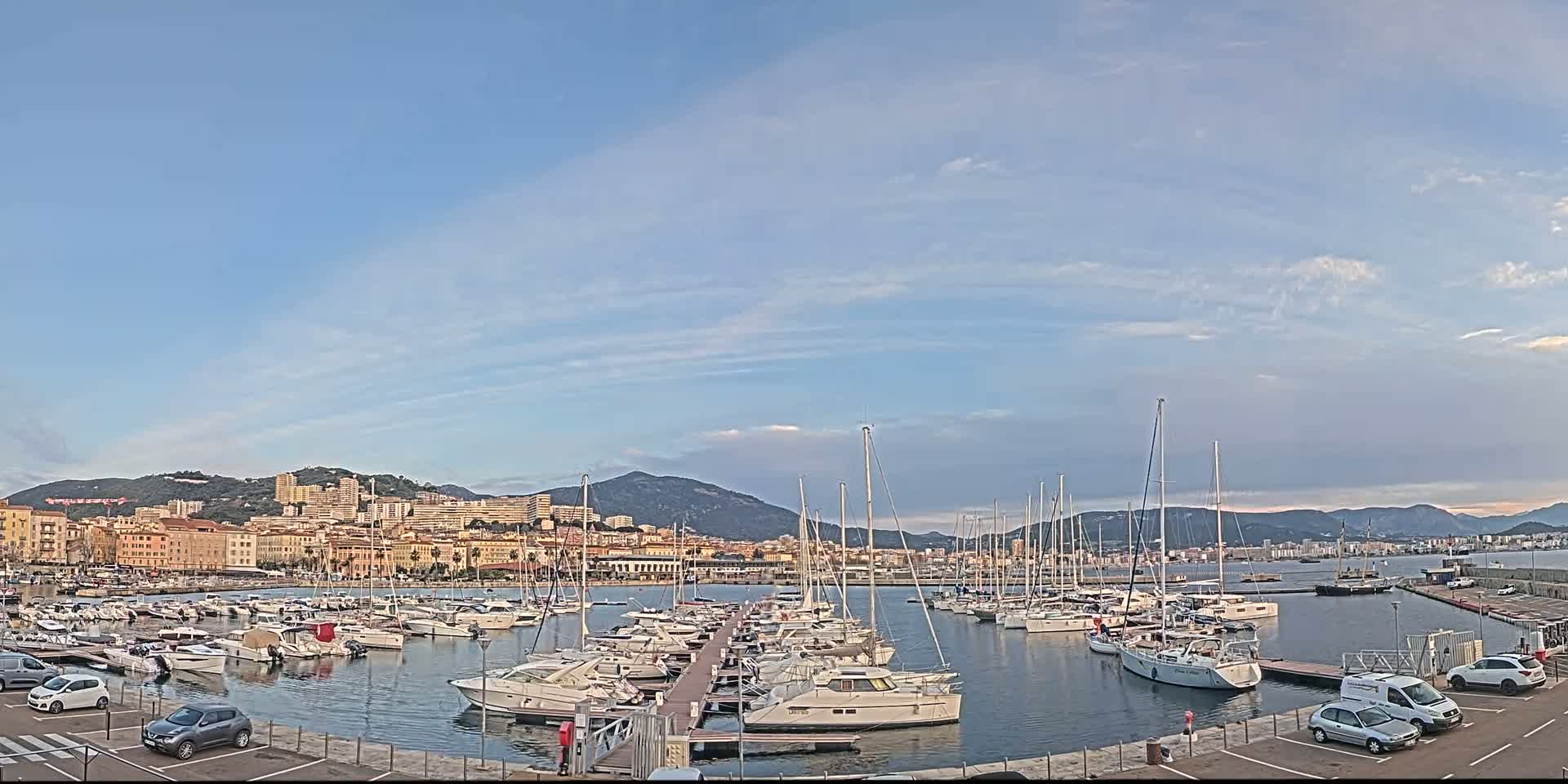 A marina filled with numerous boats is situated in front of a city nestled against a mountain range under a mostly clear sky.
