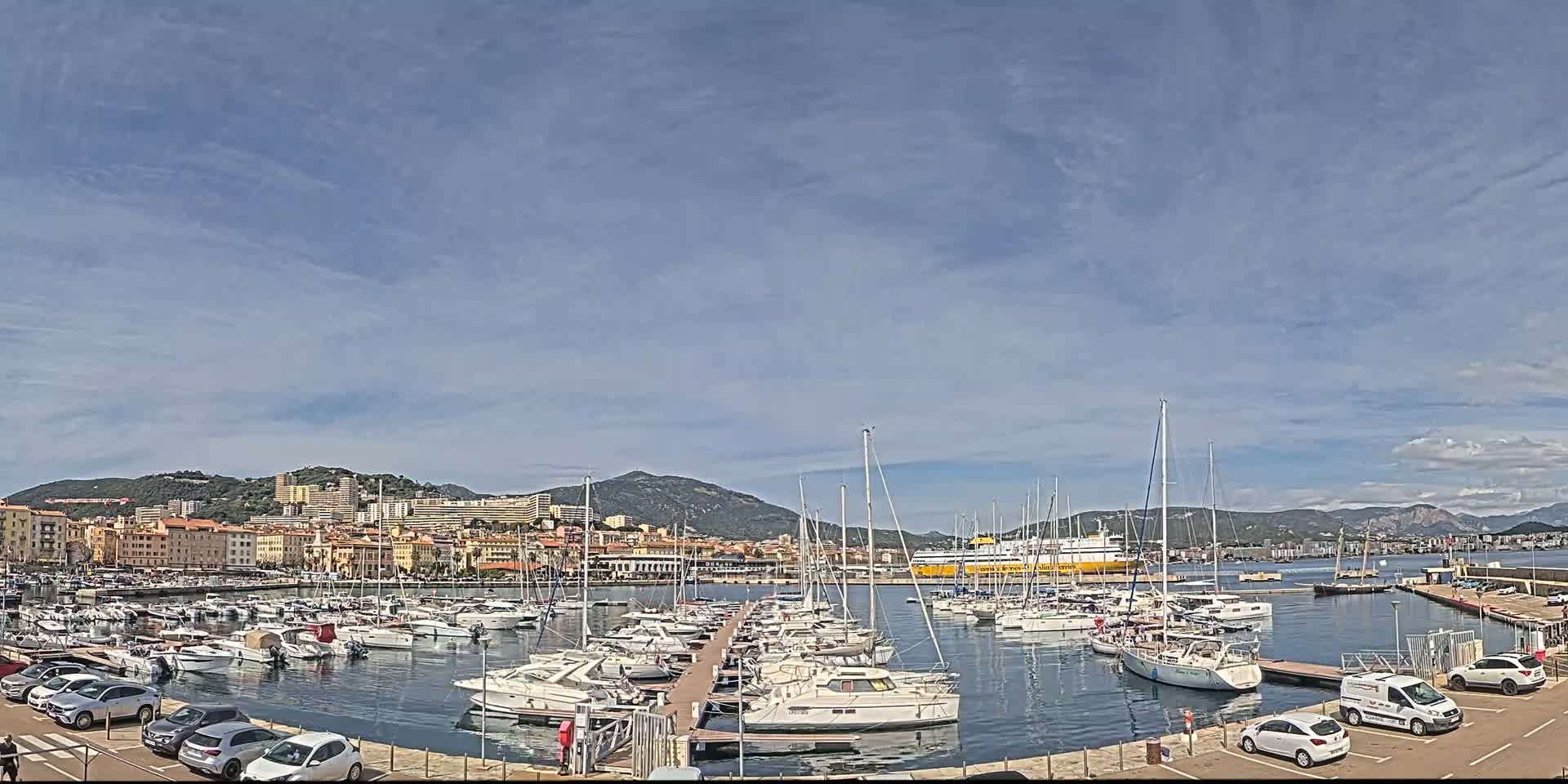 A harbor filled with numerous sailboats and motorboats is situated in front of a city nestled against a mountain range under a mostly clear sky.