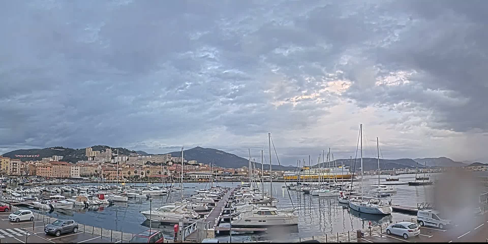 A harbor filled with numerous yachts and boats is situated under a cloudy sky, with a city and mountains in the background.