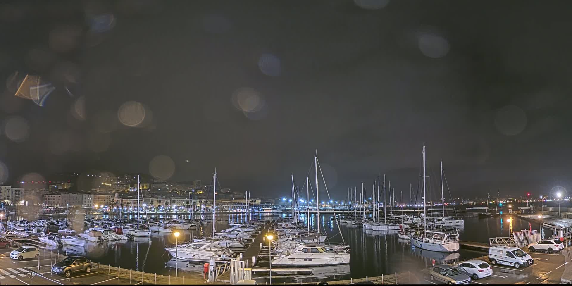 A nighttime view of a marina filled with sailboats and yachts under a dark, rainy sky.