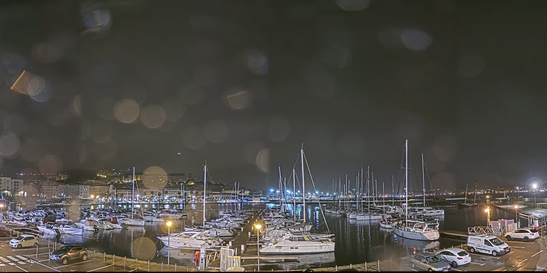 A nighttime view of a marina filled with boats during a light rain, with city lights visible in the background.