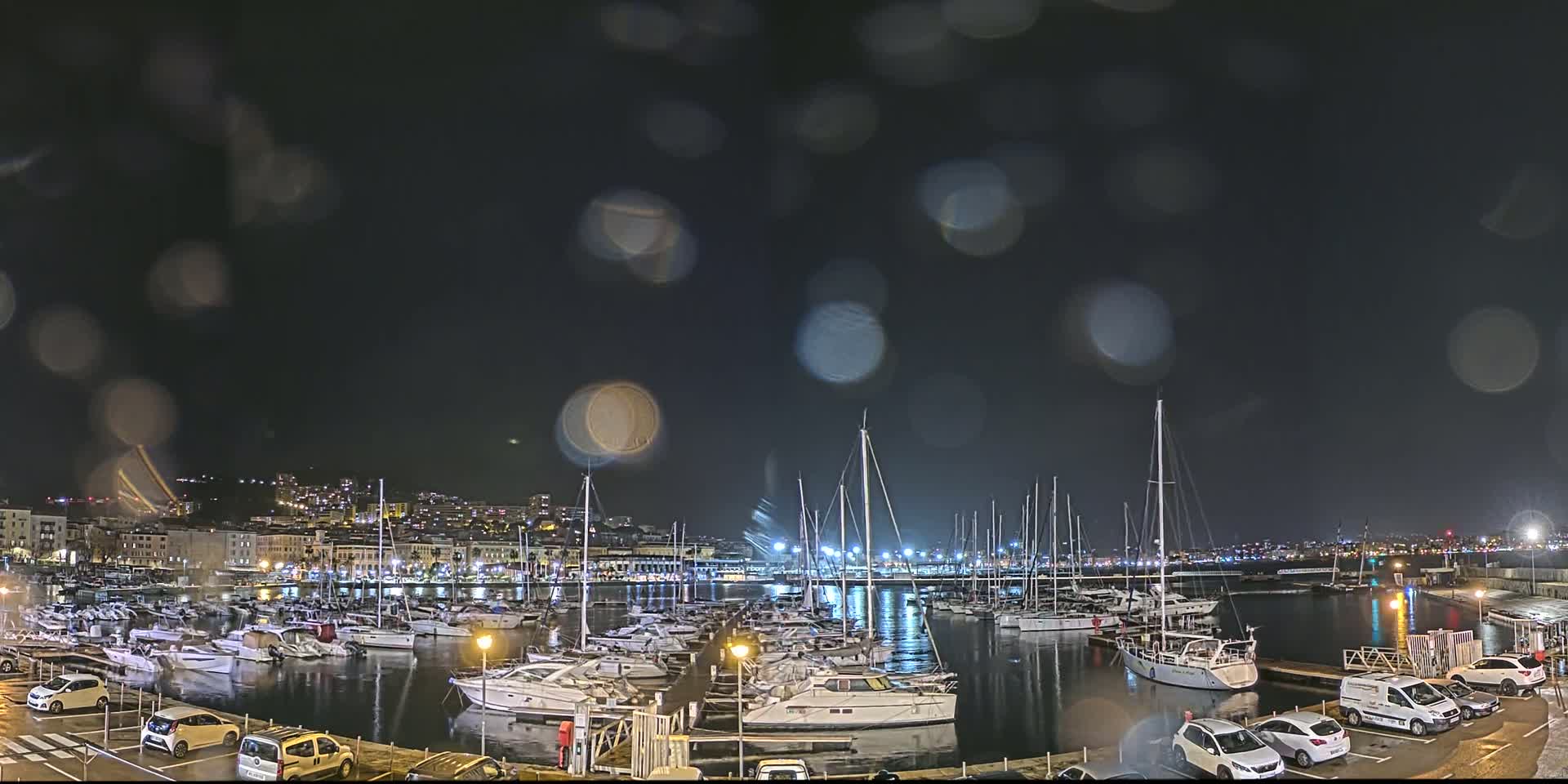 A nighttime view of a marina filled with boats during a rain shower, with a city skyline in the background.